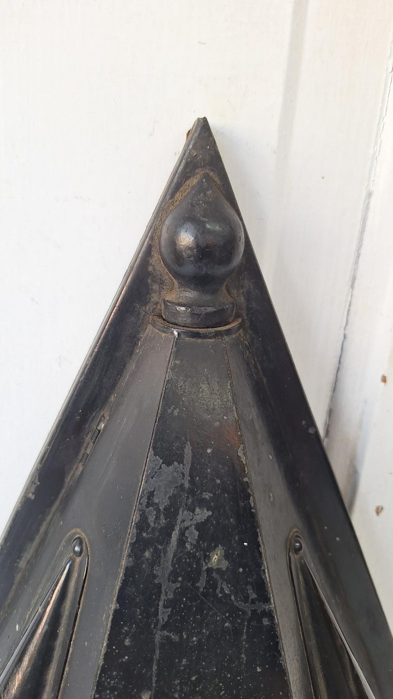 May include: A triangular black metal object with a pointed top and a central spherical finial. The surface shows signs of wear with visible paint chipping and texture. The structure features angled sides and a dark, weathered finish throughout.
