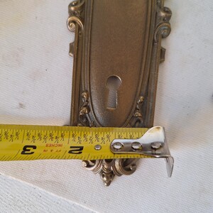 Puede incluir: Placa de puerta antigua de lat&oacute;n ornamentada con ojo de cerradura y adornos de volutas. La placa de la puerta mide aproximadamente 7,6 cm de largo. El lat&oacute;n tiene una p&aacute;tina c&aacute;lida y envejecida, y el dise&ntilde;o es sim&eacute;trico.