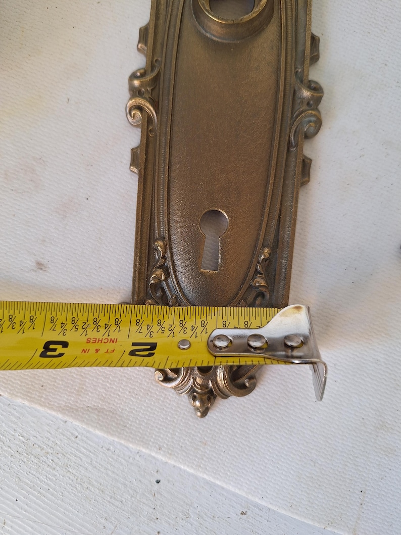 Puede incluir: Placa de puerta antigua de lat&oacute;n ornamentada con ojo de cerradura y adornos de volutas. La placa de la puerta mide aproximadamente 7,6 cm de largo. El lat&oacute;n tiene una p&aacute;tina c&aacute;lida y envejecida, y el dise&ntilde;o es sim&eacute;trico.