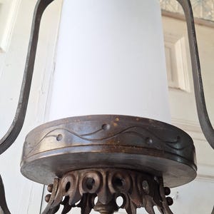 K&ouml;nnte beinhalten: Ein antiker Kronleuchter mit einem zylindrischen Schirm aus mattiertem Glas. Der Metallrahmen hat verzierte Schn&ouml;rkel und eine dunkle, gealterte Oberfl&auml;che. Die Leuchte wird an einer Kette aufgeh&auml;ngt und ist f&uuml;r die Deckenbeleuchtung konzipiert.