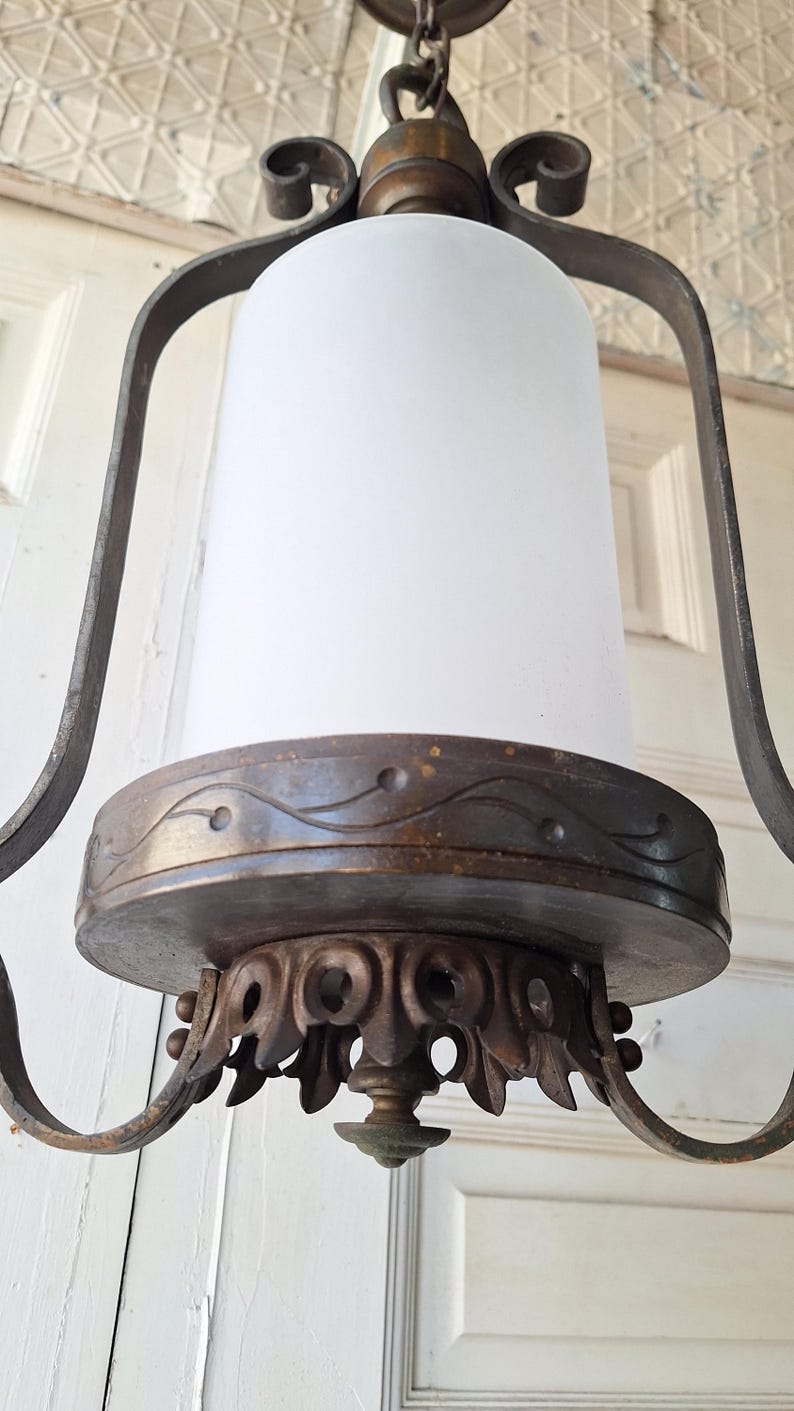K&ouml;nnte beinhalten: Ein antiker Kronleuchter mit einem zylindrischen Schirm aus mattiertem Glas. Der Metallrahmen hat verzierte Schn&ouml;rkel und eine dunkle, gealterte Oberfl&auml;che. Die Leuchte wird an einer Kette aufgeh&auml;ngt und ist f&uuml;r die Deckenbeleuchtung konzipiert.