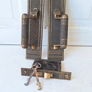 May include: Antique door hardware set featuring two long, rectangular plates with ornate detailing and gold accents. Includes matching handles, a lock, and two keys. The set is made of dark metal with intricate patterns.
