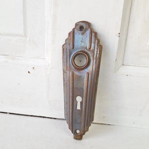 May include: An antique, Art Deco-style door plate with a keyhole and a circular opening. The metal plate has a weathered, aged appearance with vertical lines and a faded blue and brown color scheme. The door plate is mounted on a white door.