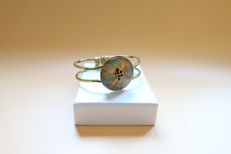 May include: A gold metal bangle bracelet with a round, colorful button featuring a floral design and black accents.