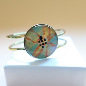 May include: A gold metal bangle bracelet with a round, blue, orange, and yellow floral design with black accents.