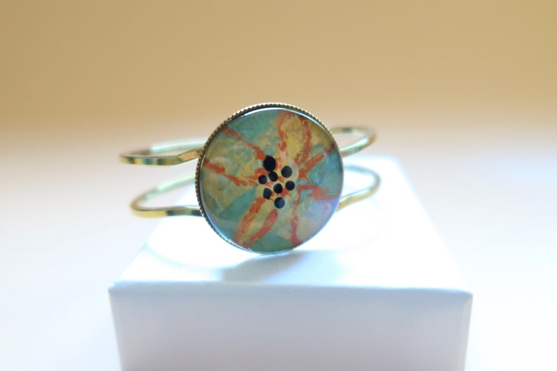 May include: A gold metal bangle bracelet with a round, blue, orange, and yellow floral design with black accents.