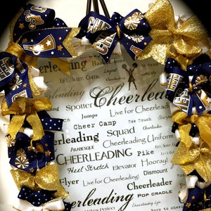 May include: A white wreath decorated with gold and navy blue cheerleading bows. The wreath has a white sign with the words "Cheerleading" and various cheerleading terms.