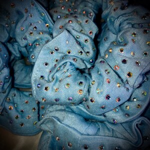May include: Close-up of several light blue scrunchies adorned with iridescent rhinestones. The fabric appears to be a soft denim-like material. The rhinestones are scattered across the surface, catching the light and creating a sparkling effect. These hair accessories are perfect for adding a touch of glamour.