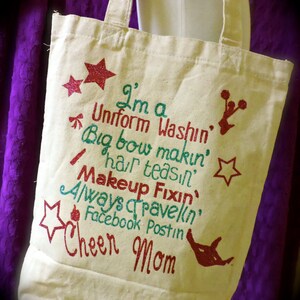 May include: A white canvas tote bag with a glittery design that says "I'm a Uniform Washin', Big bow makin', hair teasin', Makeup Fixin', Always Travelin', Facebook Postin' Cheer Mom".