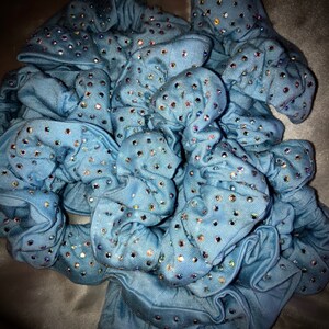 May include: A collection of light blue scrunchies, each adorned with numerous iridescent rhinestones. The scrunchies are made of a soft fabric and are bunched together, creating a textured appearance. The rhinestones sparkle, adding a touch of glamour.
