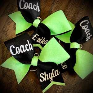 Op de afbeelding: Vier zwarte en groene cheer bows met glitter. De bows zijn versierd met witte tekst die "Coach" en de namen "Erika", "Shyla" en "Coach" zegt.