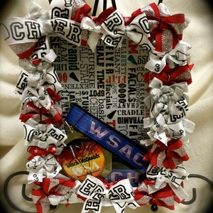 May include: A white and red cheer bow with silver glitter, a blue lanyard with the text "WSAC" in white, and a round, gold-colored medal with the text "Beach Nationals" in black. The bow is decorated with white and red ribbons and silver glitter. The lanyard is attached to the medal with a silver ring. The medal is surrounded by a white and red cheer bow with silver glitter.