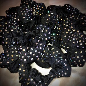 May include: A collection of black scrunchies, each adorned with numerous iridescent rhinestones. The scrunchies are made of a soft, gathered fabric, creating a textured appearance. The rhinestones sparkle, adding a touch of glamour to the hair accessories.