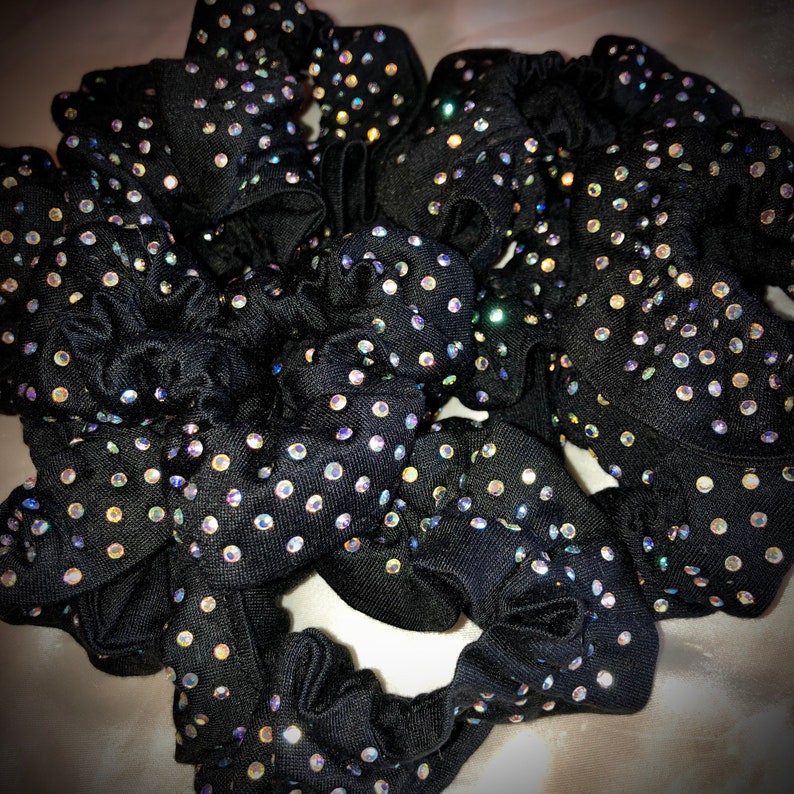 May include: A collection of black scrunchies, each adorned with numerous iridescent rhinestones. The scrunchies are made of a soft, gathered fabric, creating a textured appearance. The rhinestones sparkle, adding a touch of glamour to the hair accessories.