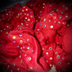 May include: Close-up of several red scrunchies adorned with iridescent rhinestones. The fabric appears to be a textured knit, and the scrunchies are bunched together, creating a layered effect. The rhinestones catch the light, adding sparkle.