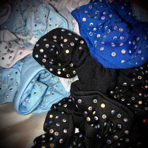 May include: A collection of scrunchies in various colors, including black, blue, and light blue. Each scrunchie is adorned with numerous sparkling rhinestones, creating a glamorous and eye-catching effect. The scrunchies are made of a soft fabric.
