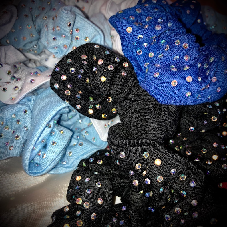 May include: A collection of scrunchies in various colors, including black, blue, and light blue. Each scrunchie is adorned with numerous sparkling rhinestones, creating a glamorous and eye-catching effect. The scrunchies are made of a soft fabric.