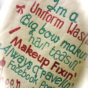 May include: A beige canvas tote bag with the text "I'm a Uniform Was Big bow makin hair teasin Fixin Makeup Always travelin' Facebook Postin'" in red and green glitter.