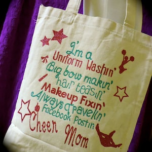 May include: A white canvas tote bag with a glittery design that says "I'm a Uniform Washin', Big Bow Makin', Hair Teas'n, Makeup Fixin', Always Travelin', Facebook Postin' Cheer Mom".