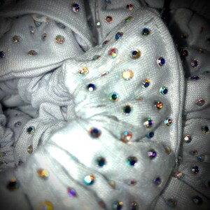 May include: Close-up of a white scrunchie adorned with numerous iridescent rhinestones. The fabric is soft and gathered, creating a textured appearance. The rhinestones reflect various colors, adding sparkle and glamour to the hair accessory.