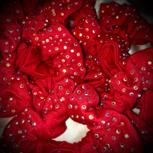 May include: A collection of red scrunchies, each adorned with numerous iridescent rhinestones. The scrunchies are made of a soft fabric and are bunched together, creating a textured and visually appealing display. The rhinestones sparkle, adding a touch of glamour.