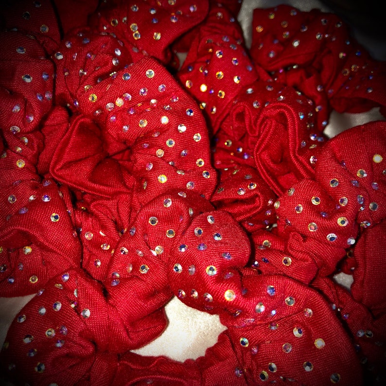 May include: A collection of red scrunchies, each adorned with numerous iridescent rhinestones. The scrunchies are made of a soft fabric and are bunched together, creating a textured and visually appealing display. The rhinestones sparkle, adding a touch of glamour.