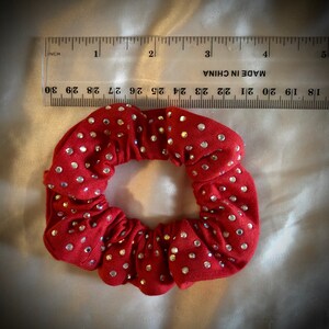 May include: A red scrunchie adorned with numerous sparkling rhinestones. The scrunchie is approximately 2 inches in diameter. The fabric is gathered, creating a textured appearance. A ruler is placed next to the scrunchie.