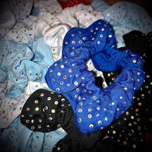 May include: A collection of fabric scrunchies in various colors, including blue, black, white, and light blue. Each scrunchie is adorned with numerous iridescent rhinestones, creating a sparkling effect. The scrunchies are bunched together, showcasing their texture and design.