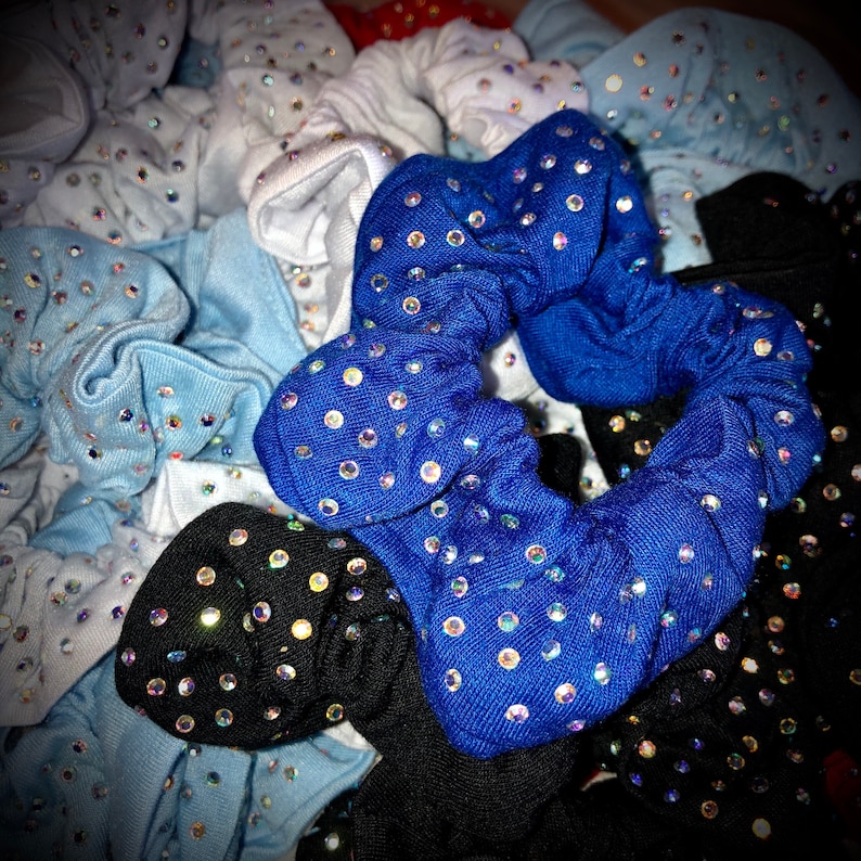 May include: A collection of fabric scrunchies in various colors, including blue, black, white, and light blue. Each scrunchie is adorned with numerous iridescent rhinestones, creating a sparkling effect. The scrunchies are bunched together, showcasing their texture and design.