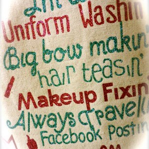 May include: A beige canvas tote bag with the text "I'm a Uniform Washin Big bow makin hair teasin Makeup Fixin Always Travel Facebook Postin" in green and red glitter.