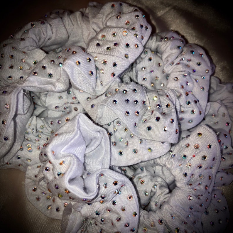 May include: A pile of light gray scrunchies, each adorned with numerous iridescent rhinestones. The scrunchies are made of a soft fabric and appear to be bunched together. The rhinestones catch the light, creating a sparkling effect.