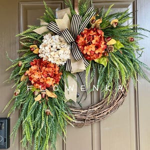 May include: A decorative wreath featuring a natural grapevine base adorned with green foliage, cream and burnt orange hydrangea blossoms, small brown acorns, and a large central bow made of tan burlap and black and white striped fabric.