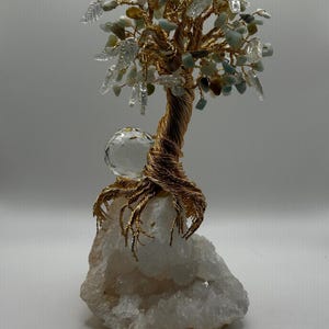 May include: A gold wire wrapped tree sculpture with green and clear crystals and a clear crystal ball at the base. The tree is rooted in a white crystal cluster.