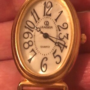 May include: A gold-toned oval-shaped watch with a white face and black Roman numeral hour markers. The watch face also has a black minute hand, a black hour hand, and a black second hand. The watch face also has the words "Leader Quartz" printed on it.