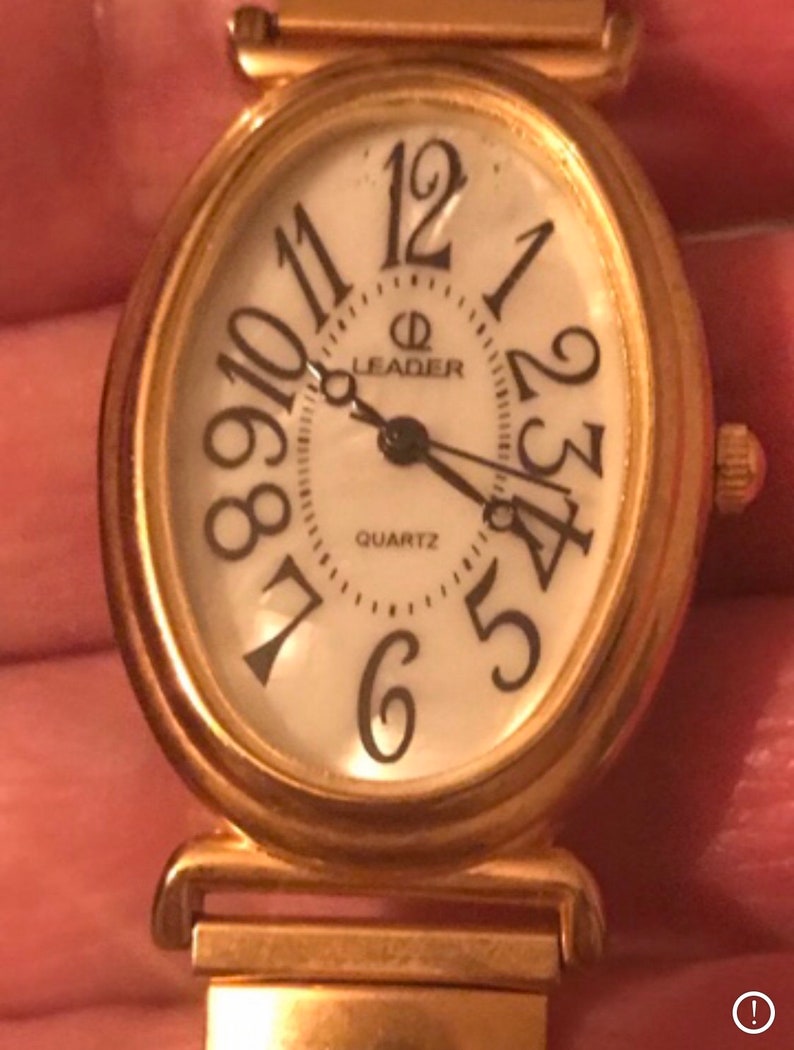 May include: A gold-toned oval-shaped watch with a white face and black Roman numeral hour markers. The watch face also has a black minute hand, a black hour hand, and a black second hand. The watch face also has the words "Leader Quartz" printed on it.