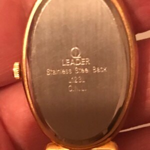 May include: Gold-tone oval watch with a stainless steel back. The watch face is blank and the back of the watch has the text "Leader Stainless Steel Back -123- C.A.L."