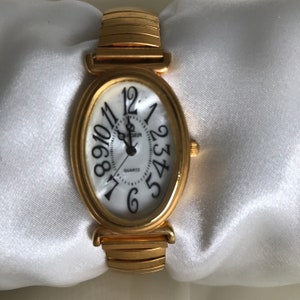 May include: A gold-tone oval-shaped watch with a white face and black Roman numeral hour markers. The watch has a gold-tone metal band with a clasp closure. The watch face also has the word "Quartz" and the brand name "Cadger" printed on it.