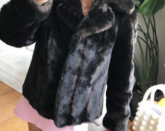 May include: A dark brown faux fur coat with a classic collar and long sleeves. The coat has a slightly textured appearance and a luxurious look. The coat is worn over a pink skirt.