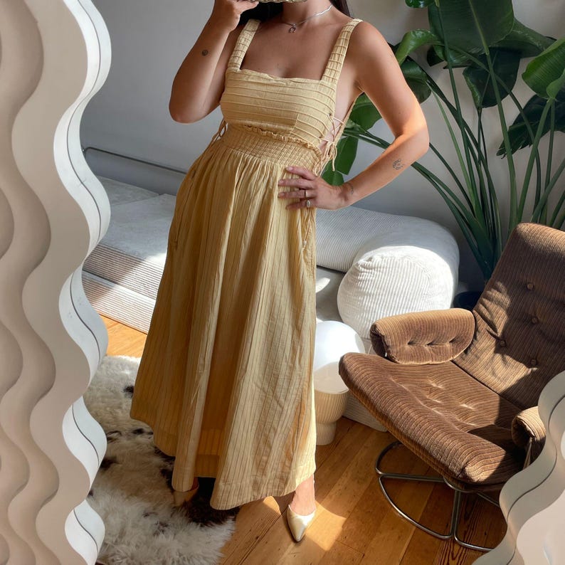 Puede incluir: Un vestido midi amarillo claro con finas rayas verticales negras. El vestido presenta tirantes gruesos, un corpi&ntilde;o ajustado y una falda fluida. El vestido tiene la cintura ce&ntilde;ida y detalles de cordones en los laterales. La modelo lleva tacones dorados.