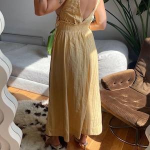 Puede incluir: Un vestido largo amarillo claro con espalda baja y detalles de lazo. El vestido tiene una textura acanalada vertical y una falda fluida. La modelo lleva tacones de color claro. El vestido es adecuado para ocasiones informales o semiformales.