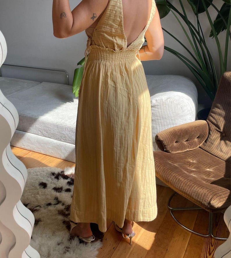 Puede incluir: Un vestido largo amarillo claro con espalda baja y detalles de lazo. El vestido tiene una textura acanalada vertical y una falda fluida. La modelo lleva tacones de color claro. El vestido es adecuado para ocasiones informales o semiformales.