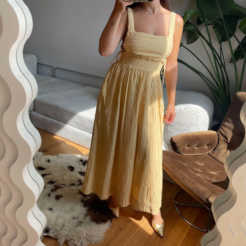 Puede incluir: Un vestido de verano amarillo claro con rayas verticales. El vestido tiene un escote cuadrado, tirantes anchos y un corpi&ntilde;o ajustado con cintura fruncida. La falda llega hasta la altura de la pantorrilla. El vestido se combina con tacones dorados.