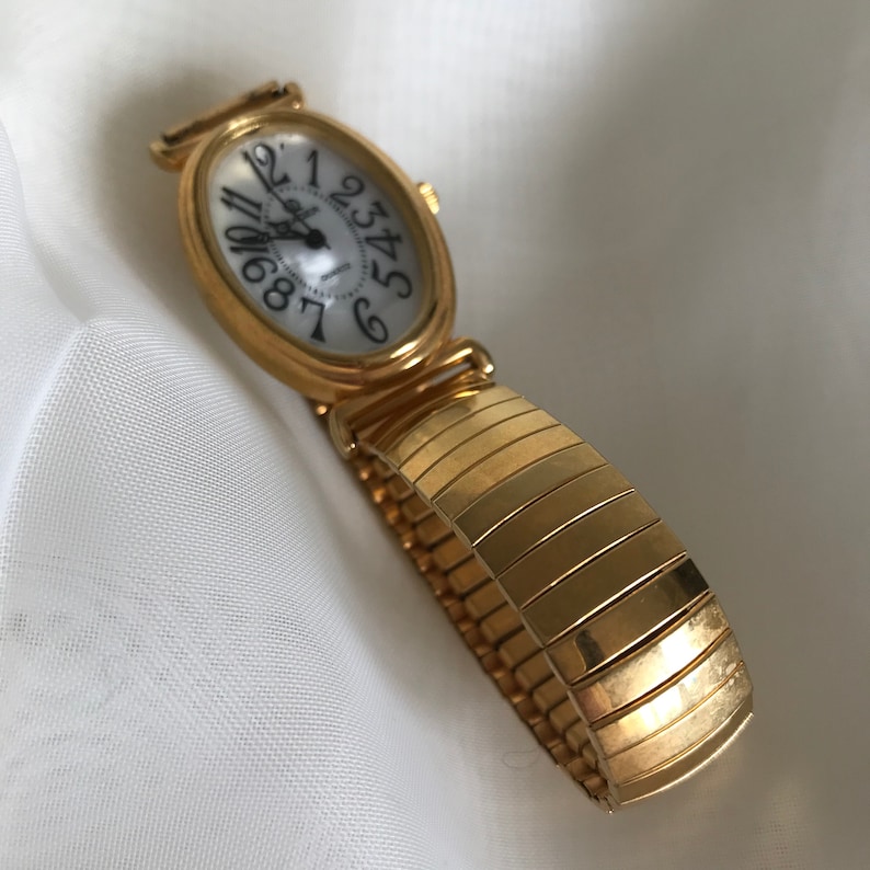 May include: A gold-toned oval-shaped wristwatch with a white face and black Roman numeral hour markers. The watch has a gold-toned expandable band.