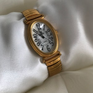 May include: A gold-tone oval-shaped watch with a white face and black Roman numeral hour markers. The watch has a gold-tone metal band.