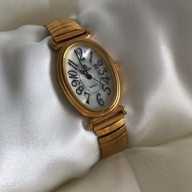 May include: A gold-tone oval-shaped watch with a white face and black Roman numeral hour markers. The watch has a gold-tone metal band.
