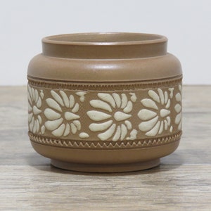 May include: A brown ceramic jar with a decorative floral pattern in cream. The jar has a cylindrical shape with a slightly flared rim and a base. The floral design is embossed around the middle of the jar.