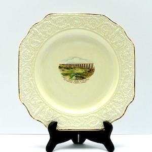 May include: Cream-colored, octagonal decorative plate with a gold rim and a central image of a landscape. The plate has a raised floral pattern around the edge and is displayed on a black stand. The text on the plate reads "Lethbridge Viaduct, Alberta, Canada."