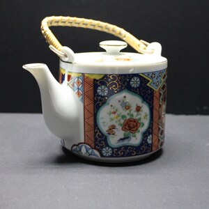 May include: A white porcelain teapot featuring a cylindrical body with a decorative pattern of dark blue, orange, and gold geometric shapes and floral motifs. It includes a curved spout, a matching lid, and a woven light-colored bamboo handle.
