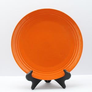 May include: A vibrant orange ceramic plate with concentric rings near the rim. The plate is displayed on a black, three-legged stand against a white backdrop. This decorative plate is suitable for serving food.