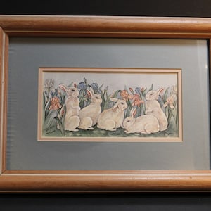 May include: Framed watercolor painting of six white rabbits sitting in a field of green grass and colorful flowers. The rabbits are all facing forward and have their ears perked up. The flowers are a mix of pink, purple, and blue.